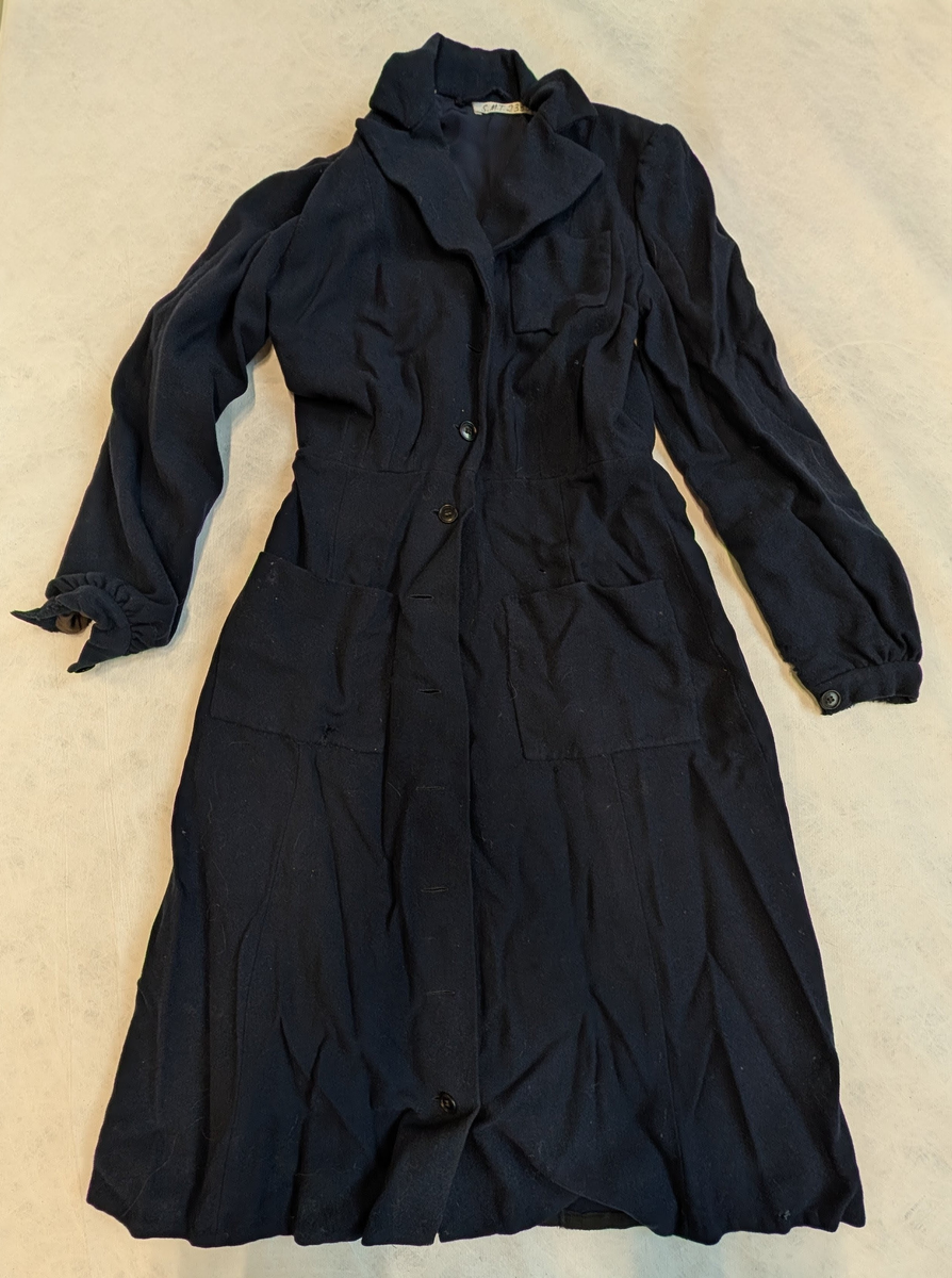 Kjolen er sydd sammen i livet av en overdel med lange ermer og et utskåret skjørt. Overdelen er helfôret med “crem de skin” og et rayonstoff. Ermene er rynket ved håndleddet og har linning som kneppes med én knapp. Hele kjolen kneppes foran med ovale knapper. Den har krage med slag, én lomme på venstre side av brystet og to større lommer på skjørtet, én på hver side. Overdelen er rynket ned mot skjørtet. Form: frakkekjole med knepping foran.