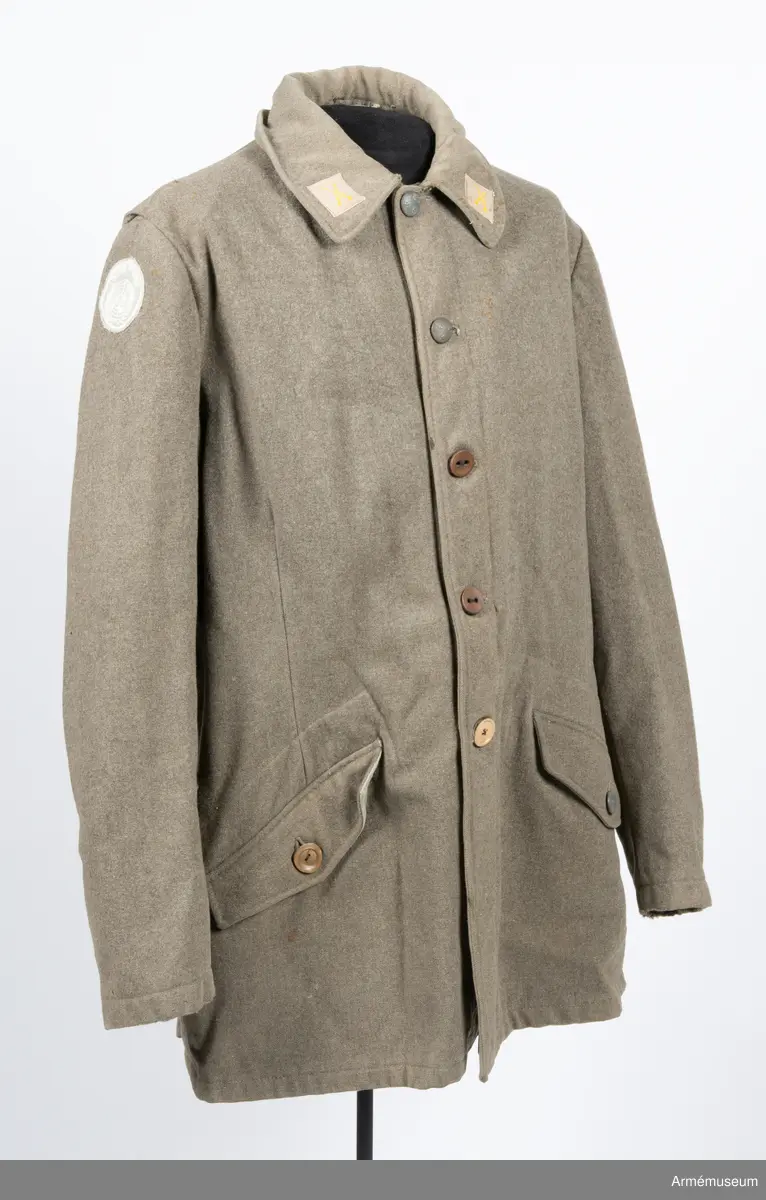 Gråbrungrön vapenrock med bälgfickor bak.

Rocken är sydd av så kallad gråbrungrönt kläde och är fodrad upptill i bålen med gråbrunt bomullstyg. Ärmarna är ofodrade. Den är konstruerad med helt framstycke med midjeinsnitt, sidstycke bak, samt bakstycke med mitt bak-söm. På varje sida av bakstycket finns ett lagt veck som är nedsydd ner till midjan. Ärmarna är tvåsömsärmar. Under ärmarna finns ovala ärmspjäll av fodertyget. Kragen är nedliggande med kragspeglar med två korslagda svärd i gult.
Två stycken snedställda fickor nedtill på framsidan samt två horisontella bälgfickor baktill. Alla fickorna har lock. Knäppningen mitt fram har haft grå metallknappar med tre kronor på räfflad botten. Bara två av dessa finns kvar och har ersatts med diverse civila plastknappar. Vänster framficka och de två bakfickorna har originalknapparna kvar.
Plagget är märkt på insidan med en skrödderietikett med text i vitt på grå botten: "Sydd av Söderbergs Upsala".
På vänster ärm är fastsytt ett runt märke för UN, ursprungligen i vitt på blå botten men numera bleknat. På höger ärm är "Sweden" samt ett märke med Tre kronor i gult på blå botten slarvigt fastsytt.
I vänster framstycke finns på fodersidan en liten innerficka samt en större med lock. Baktill sitter i nederkant en spänntamp avsedd att kunna knäppas mellan benen. Denna är numera nedsydd.
I nacken sitter en hank av fodertyget.