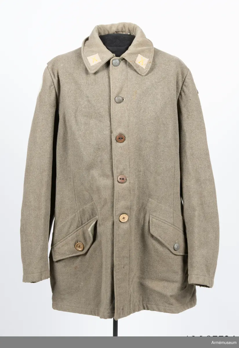 Gråbrungrön vapenrock med bälgfickor bak.

Rocken är sydd av så kallad gråbrungrönt kläde och är fodrad upptill i bålen med gråbrunt bomullstyg. Ärmarna är ofodrade. Den är konstruerad med helt framstycke med midjeinsnitt, sidstycke bak, samt bakstycke med mitt bak-söm. På varje sida av bakstycket finns ett lagt veck som är nedsydd ner till midjan. Ärmarna är tvåsömsärmar. Under ärmarna finns ovala ärmspjäll av fodertyget. Kragen är nedliggande med kragspeglar med två korslagda svärd i gult.
Två stycken snedställda fickor nedtill på framsidan samt två horisontella bälgfickor baktill. Alla fickorna har lock. Knäppningen mitt fram har haft grå metallknappar med tre kronor på räfflad botten. Bara två av dessa finns kvar och har ersatts med diverse civila plastknappar. Vänster framficka och de två bakfickorna har originalknapparna kvar.
Plagget är märkt på insidan med en skrödderietikett med text i vitt på grå botten: "Sydd av Söderbergs Upsala".
På vänster ärm är fastsytt ett runt märke för UN, ursprungligen i vitt på blå botten men numera bleknat. På höger ärm är "Sweden" samt ett märke med Tre kronor i gult på blå botten slarvigt fastsytt.
I vänster framstycke finns på fodersidan en liten innerficka samt en större med lock. Baktill sitter i nederkant en spänntamp avsedd att kunna knäppas mellan benen. Denna är numera nedsydd.
I nacken sitter en hank av fodertyget.