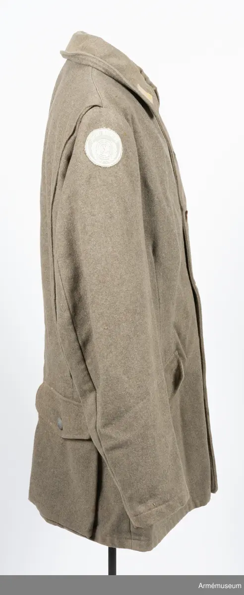 Gråbrungrön vapenrock med bälgfickor bak.

Rocken är sydd av så kallad gråbrungrönt kläde och är fodrad upptill i bålen med gråbrunt bomullstyg. Ärmarna är ofodrade. Den är konstruerad med helt framstycke med midjeinsnitt, sidstycke bak, samt bakstycke med mitt bak-söm. På varje sida av bakstycket finns ett lagt veck som är nedsydd ner till midjan. Ärmarna är tvåsömsärmar. Under ärmarna finns ovala ärmspjäll av fodertyget. Kragen är nedliggande med kragspeglar med två korslagda svärd i gult.
Två stycken snedställda fickor nedtill på framsidan samt två horisontella bälgfickor baktill. Alla fickorna har lock. Knäppningen mitt fram har haft grå metallknappar med tre kronor på räfflad botten. Bara två av dessa finns kvar och har ersatts med diverse civila plastknappar. Vänster framficka och de två bakfickorna har originalknapparna kvar.
Plagget är märkt på insidan med en skrödderietikett med text i vitt på grå botten: "Sydd av Söderbergs Upsala".
På vänster ärm är fastsytt ett runt märke för UN, ursprungligen i vitt på blå botten men numera bleknat. På höger ärm är "Sweden" samt ett märke med Tre kronor i gult på blå botten slarvigt fastsytt.
I vänster framstycke finns på fodersidan en liten innerficka samt en större med lock. Baktill sitter i nederkant en spänntamp avsedd att kunna knäppas mellan benen. Denna är numera nedsydd.
I nacken sitter en hank av fodertyget.