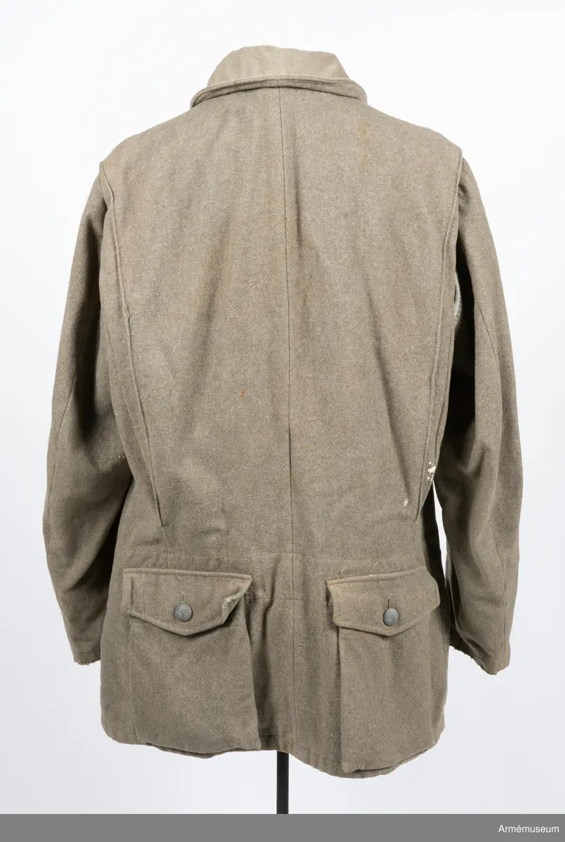 Gråbrungrön vapenrock med bälgfickor bak.

Rocken är sydd av så kallad gråbrungrönt kläde och är fodrad upptill i bålen med gråbrunt bomullstyg. Ärmarna är ofodrade. Den är konstruerad med helt framstycke med midjeinsnitt, sidstycke bak, samt bakstycke med mitt bak-söm. På varje sida av bakstycket finns ett lagt veck som är nedsydd ner till midjan. Ärmarna är tvåsömsärmar. Under ärmarna finns ovala ärmspjäll av fodertyget. Kragen är nedliggande med kragspeglar med två korslagda svärd i gult.
Två stycken snedställda fickor nedtill på framsidan samt två horisontella bälgfickor baktill. Alla fickorna har lock. Knäppningen mitt fram har haft grå metallknappar med tre kronor på räfflad botten. Bara två av dessa finns kvar och har ersatts med diverse civila plastknappar. Vänster framficka och de två bakfickorna har originalknapparna kvar.
Plagget är märkt på insidan med en skrödderietikett med text i vitt på grå botten: "Sydd av Söderbergs Upsala".
På vänster ärm är fastsytt ett runt märke för UN, ursprungligen i vitt på blå botten men numera bleknat. På höger ärm är "Sweden" samt ett märke med Tre kronor i gult på blå botten slarvigt fastsytt.
I vänster framstycke finns på fodersidan en liten innerficka samt en större med lock. Baktill sitter i nederkant en spänntamp avsedd att kunna knäppas mellan benen. Denna är numera nedsydd.
I nacken sitter en hank av fodertyget.