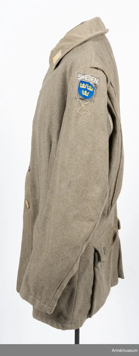 Gråbrungrön vapenrock med bälgfickor bak.

Rocken är sydd av så kallad gråbrungrönt kläde och är fodrad upptill i bålen med gråbrunt bomullstyg. Ärmarna är ofodrade. Den är konstruerad med helt framstycke med midjeinsnitt, sidstycke bak, samt bakstycke med mitt bak-söm. På varje sida av bakstycket finns ett lagt veck som är nedsydd ner till midjan. Ärmarna är tvåsömsärmar. Under ärmarna finns ovala ärmspjäll av fodertyget. Kragen är nedliggande med kragspeglar med två korslagda svärd i gult.
Två stycken snedställda fickor nedtill på framsidan samt två horisontella bälgfickor baktill. Alla fickorna har lock. Knäppningen mitt fram har haft grå metallknappar med tre kronor på räfflad botten. Bara två av dessa finns kvar och har ersatts med diverse civila plastknappar. Vänster framficka och de två bakfickorna har originalknapparna kvar.
Plagget är märkt på insidan med en skrödderietikett med text i vitt på grå botten: "Sydd av Söderbergs Upsala".
På vänster ärm är fastsytt ett runt märke för UN, ursprungligen i vitt på blå botten men numera bleknat. På höger ärm är "Sweden" samt ett märke med Tre kronor i gult på blå botten slarvigt fastsytt.
I vänster framstycke finns på fodersidan en liten innerficka samt en större med lock. Baktill sitter i nederkant en spänntamp avsedd att kunna knäppas mellan benen. Denna är numera nedsydd.
I nacken sitter en hank av fodertyget.