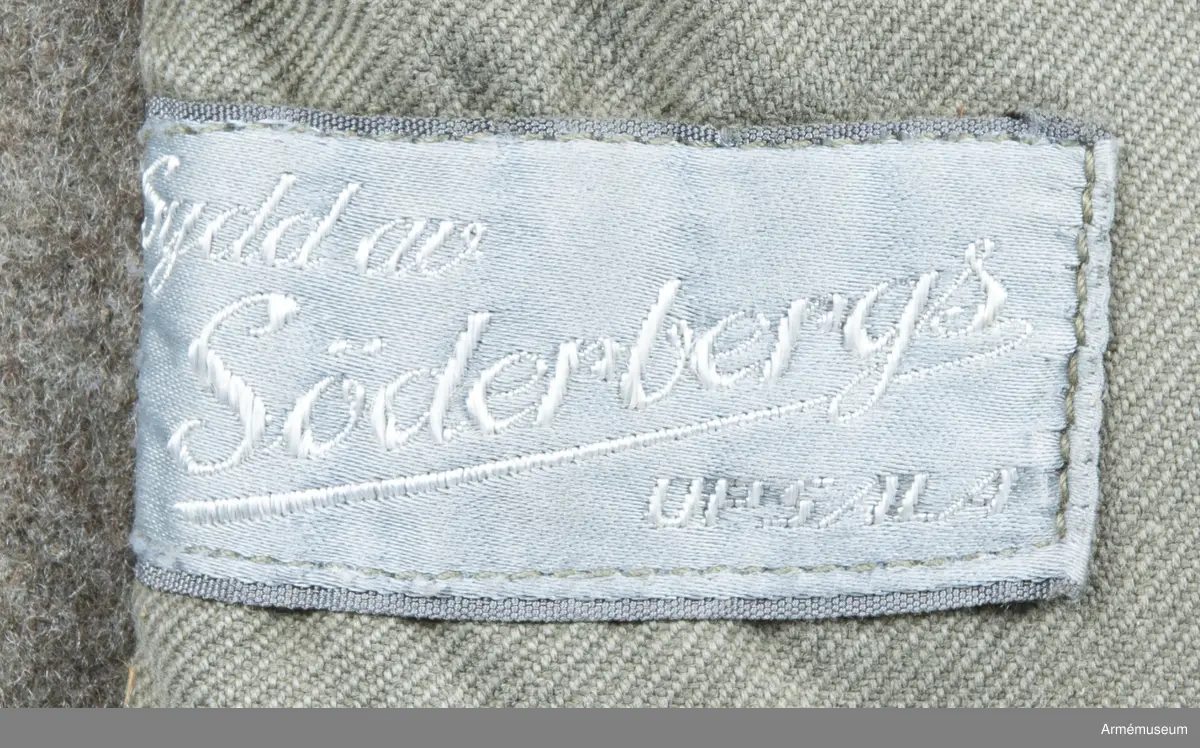 Gråbrungrön vapenrock med bälgfickor bak.

Rocken är sydd av så kallad gråbrungrönt kläde och är fodrad upptill i bålen med gråbrunt bomullstyg. Ärmarna är ofodrade. Den är konstruerad med helt framstycke med midjeinsnitt, sidstycke bak, samt bakstycke med mitt bak-söm. På varje sida av bakstycket finns ett lagt veck som är nedsydd ner till midjan. Ärmarna är tvåsömsärmar. Under ärmarna finns ovala ärmspjäll av fodertyget. Kragen är nedliggande med kragspeglar med två korslagda svärd i gult.
Två stycken snedställda fickor nedtill på framsidan samt två horisontella bälgfickor baktill. Alla fickorna har lock. Knäppningen mitt fram har haft grå metallknappar med tre kronor på räfflad botten. Bara två av dessa finns kvar och har ersatts med diverse civila plastknappar. Vänster framficka och de två bakfickorna har originalknapparna kvar.
Plagget är märkt på insidan med en skrödderietikett med text i vitt på grå botten: "Sydd av Söderbergs Upsala".
På vänster ärm är fastsytt ett runt märke för UN, ursprungligen i vitt på blå botten men numera bleknat. På höger ärm är "Sweden" samt ett märke med Tre kronor i gult på blå botten slarvigt fastsytt.
I vänster framstycke finns på fodersidan en liten innerficka samt en större med lock. Baktill sitter i nederkant en spänntamp avsedd att kunna knäppas mellan benen. Denna är numera nedsydd.
I nacken sitter en hank av fodertyget.