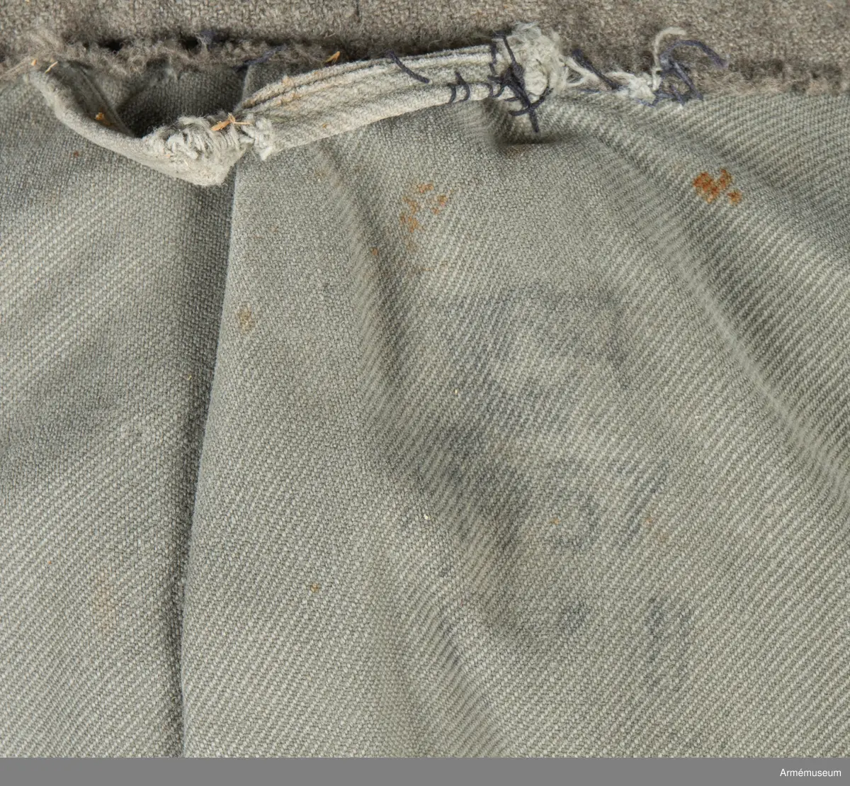 Gråbrungrön vapenrock med bälgfickor bak.

Rocken är sydd av så kallad gråbrungrönt kläde och är fodrad upptill i bålen med gråbrunt bomullstyg. Ärmarna är ofodrade. Den är konstruerad med helt framstycke med midjeinsnitt, sidstycke bak, samt bakstycke med mitt bak-söm. På varje sida av bakstycket finns ett lagt veck som är nedsydd ner till midjan. Ärmarna är tvåsömsärmar. Under ärmarna finns ovala ärmspjäll av fodertyget. Kragen är nedliggande med kragspeglar med två korslagda svärd i gult.
Två stycken snedställda fickor nedtill på framsidan samt två horisontella bälgfickor baktill. Alla fickorna har lock. Knäppningen mitt fram har haft grå metallknappar med tre kronor på räfflad botten. Bara två av dessa finns kvar och har ersatts med diverse civila plastknappar. Vänster framficka och de två bakfickorna har originalknapparna kvar.
Plagget är märkt på insidan med en skrödderietikett med text i vitt på grå botten: "Sydd av Söderbergs Upsala".
På vänster ärm är fastsytt ett runt märke för UN, ursprungligen i vitt på blå botten men numera bleknat. På höger ärm är "Sweden" samt ett märke med Tre kronor i gult på blå botten slarvigt fastsytt.
I vänster framstycke finns på fodersidan en liten innerficka samt en större med lock. Baktill sitter i nederkant en spänntamp avsedd att kunna knäppas mellan benen. Denna är numera nedsydd.
I nacken sitter en hank av fodertyget.