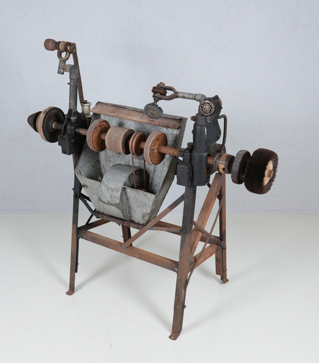 Pussemaskin for sko. Underdel i metall (4 bein). Oppå underdelen er det montert anordning i hardt tre og metall, mellom anna ein aksling i metall. Sandpapir og kostar er feste på sirkelforma delar, som er feste inn på akslingen. Under delane med sandpapir er det montert ei oppsamlingsanordning i blikk. Denne har opning ut på andre sida.