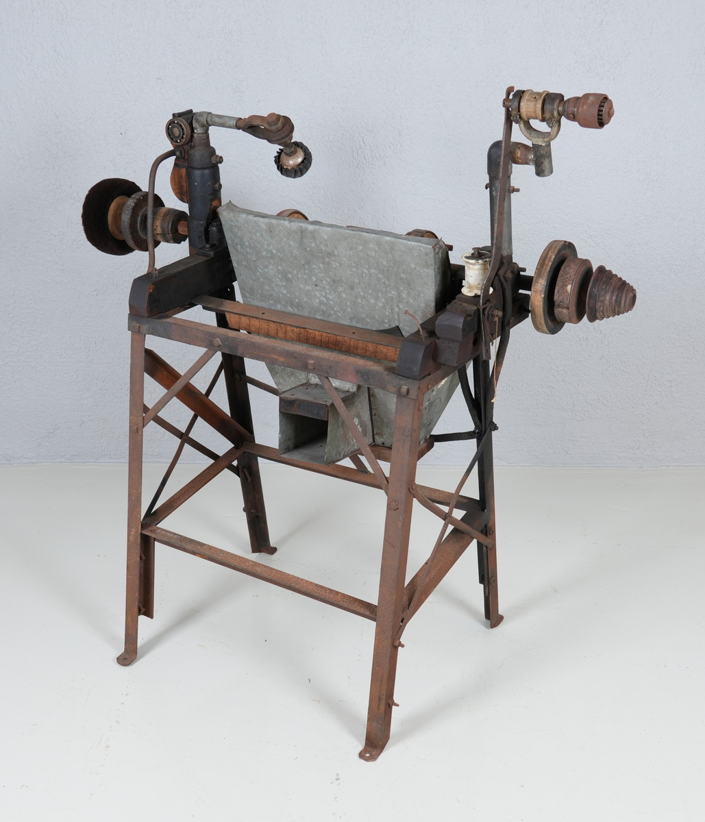 Pussemaskin for sko. Underdel i metall (4 bein). Oppå underdelen er det montert anordning i hardt tre og metall, mellom anna ein aksling i metall. Sandpapir og kostar er feste på sirkelforma delar, som er feste inn på akslingen. Under delane med sandpapir er det montert ei oppsamlingsanordning i blikk. Denne har opning ut på andre sida.