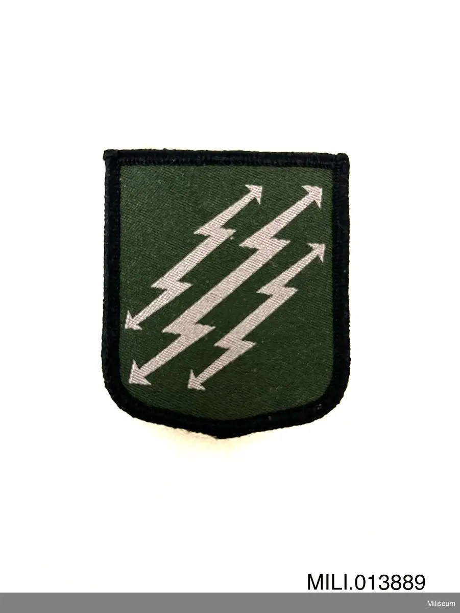 Tilläggstecken av textil med maskinvävt motiv. Med kardborrefästning på baksidan. Avsett att fästas på vapenrockens (motsv.) vänstra överärm.