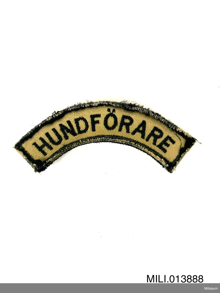 Tilläggstecken av textil med maskinbroderad text. Med kardborrefästning på baksidan. Avsett att fästas på vapenrockens (motsv.) högra överärm.