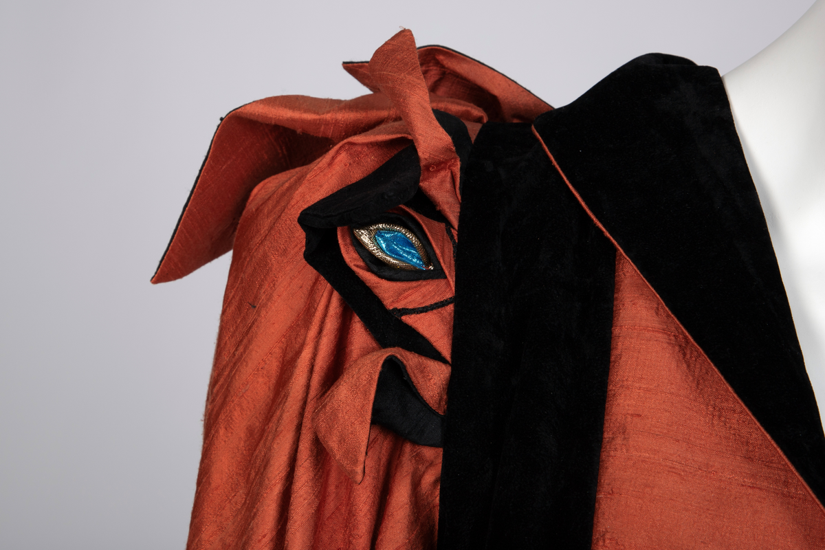 Ensemble bestående av kåpe, overdel, haremsbukse, hatt og mansjetter. Lang kåpe i rustrød råsilke med draperte ermer og detaljer i svart fløyel. Bladlignende dekor av blått og gullfarget stoff ved høyre skulder. Ermeløs overdel i svart råsilke og fløyel med svart syntetisk fôr. Overdelen går i en spiss opp ved bysten og i en spiss ned ved hoftene. Smale stropper over skuldrene. Dekorblomst i blått, gull og rustrødt i navlehøyde midt foran. Haremsbukse i svart syntetisk mønstervevd stoff med knapper kledd i rustrød råsilke nederst. Asymmetrisk hatt i svart ull- og silkeblanding, rustrød råsilke og polyesterbånd på innsiden. Mansjetter i råsilke med bomullsfôr.