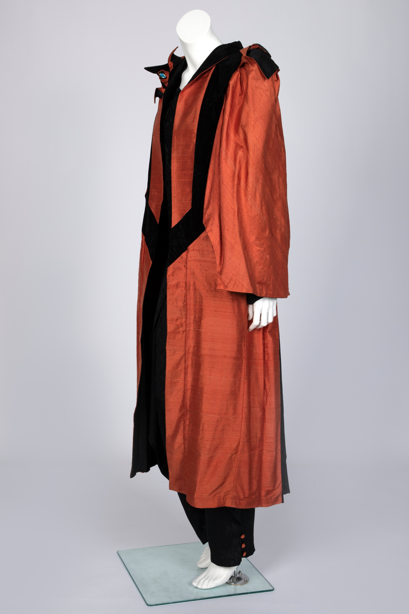 Ensemble bestående av kåpe, overdel, haremsbukse, hatt og mansjetter. Lang kåpe i rustrød råsilke med draperte ermer og detaljer i svart fløyel. Bladlignende dekor av blått og gullfarget stoff ved høyre skulder. Ermeløs overdel i svart råsilke og fløyel med svart syntetisk fôr. Overdelen går i en spiss opp ved bysten og i en spiss ned ved hoftene. Smale stropper over skuldrene. Dekorblomst i blått, gull og rustrødt i navlehøyde midt foran. Haremsbukse i svart syntetisk mønstervevd stoff med knapper kledd i rustrød råsilke nederst. Asymmetrisk hatt i svart ull- og silkeblanding, rustrød råsilke og polyesterbånd på innsiden. Mansjetter i råsilke med bomullsfôr.