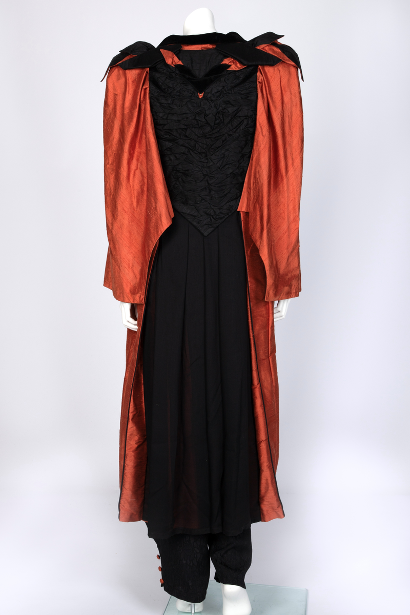 Ensemble bestående av kåpe, overdel, haremsbukse, hatt og mansjetter. Lang kåpe i rustrød råsilke med draperte ermer og detaljer i svart fløyel. Bladlignende dekor av blått og gullfarget stoff ved høyre skulder. Ermeløs overdel i svart råsilke og fløyel med svart syntetisk fôr. Overdelen går i en spiss opp ved bysten og i en spiss ned ved hoftene. Smale stropper over skuldrene. Dekorblomst i blått, gull og rustrødt i navlehøyde midt foran. Haremsbukse i svart syntetisk mønstervevd stoff med knapper kledd i rustrød råsilke nederst. Asymmetrisk hatt i svart ull- og silkeblanding, rustrød råsilke og polyesterbånd på innsiden. Mansjetter i råsilke med bomullsfôr.