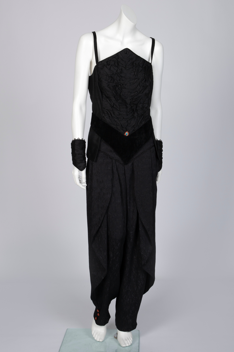 Ensemble bestående av kåpe, overdel, haremsbukse, hatt og mansjetter. Lang kåpe i rustrød råsilke med draperte ermer og detaljer i svart fløyel. Bladlignende dekor av blått og gullfarget stoff ved høyre skulder. Ermeløs overdel i svart råsilke og fløyel med svart syntetisk fôr. Overdelen går i en spiss opp ved bysten og i en spiss ned ved hoftene. Smale stropper over skuldrene. Dekorblomst i blått, gull og rustrødt i navlehøyde midt foran. Haremsbukse i svart syntetisk mønstervevd stoff med knapper kledd i rustrød råsilke nederst. Asymmetrisk hatt i svart ull- og silkeblanding, rustrød råsilke og polyesterbånd på innsiden. Mansjetter i råsilke med bomullsfôr.