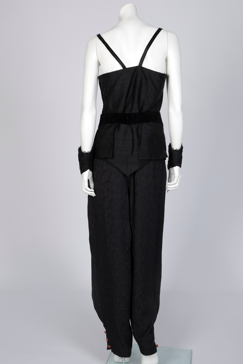 Ensemble bestående av kåpe, overdel, haremsbukse, hatt og mansjetter. Lang kåpe i rustrød råsilke med draperte ermer og detaljer i svart fløyel. Bladlignende dekor av blått og gullfarget stoff ved høyre skulder. Ermeløs overdel i svart råsilke og fløyel med svart syntetisk fôr. Overdelen går i en spiss opp ved bysten og i en spiss ned ved hoftene. Smale stropper over skuldrene. Dekorblomst i blått, gull og rustrødt i navlehøyde midt foran. Haremsbukse i svart syntetisk mønstervevd stoff med knapper kledd i rustrød råsilke nederst. Asymmetrisk hatt i svart ull- og silkeblanding, rustrød råsilke og polyesterbånd på innsiden. Mansjetter i råsilke med bomullsfôr.