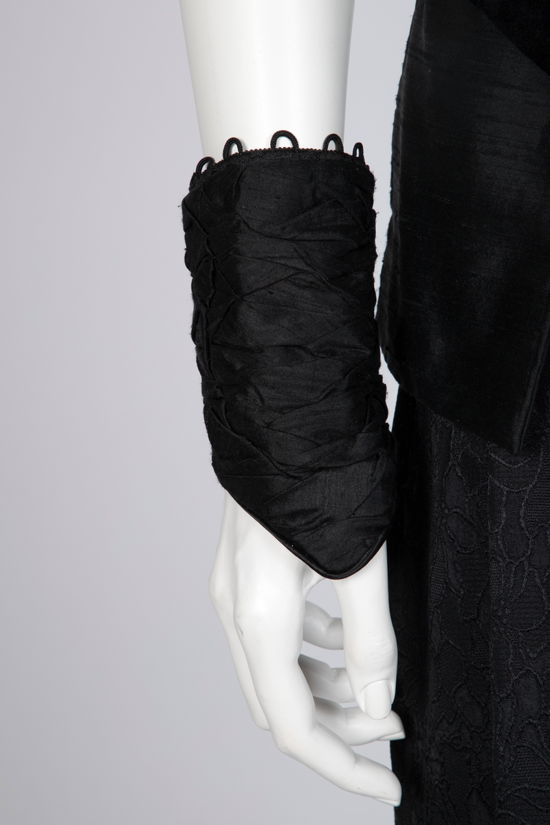 Ensemble bestående av kåpe, overdel, haremsbukse, hatt og mansjetter. Lang kåpe i rustrød råsilke med draperte ermer og detaljer i svart fløyel. Bladlignende dekor av blått og gullfarget stoff ved høyre skulder. Ermeløs overdel i svart råsilke og fløyel med svart syntetisk fôr. Overdelen går i en spiss opp ved bysten og i en spiss ned ved hoftene. Smale stropper over skuldrene. Dekorblomst i blått, gull og rustrødt i navlehøyde midt foran. Haremsbukse i svart syntetisk mønstervevd stoff med knapper kledd i rustrød råsilke nederst. Asymmetrisk hatt i svart ull- og silkeblanding, rustrød råsilke og polyesterbånd på innsiden. Mansjetter i råsilke med bomullsfôr.