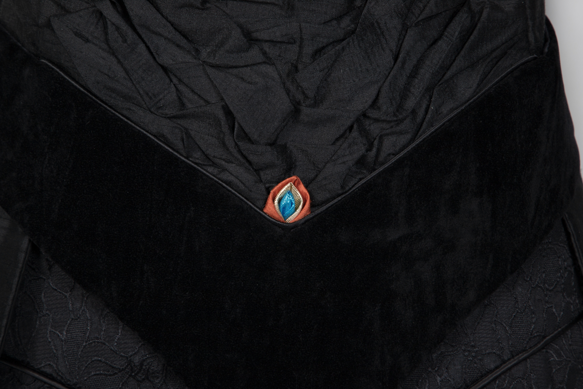 Ensemble bestående av kåpe, overdel, haremsbukse, hatt og mansjetter. Lang kåpe i rustrød råsilke med draperte ermer og detaljer i svart fløyel. Bladlignende dekor av blått og gullfarget stoff ved høyre skulder. Ermeløs overdel i svart råsilke og fløyel med svart syntetisk fôr. Overdelen går i en spiss opp ved bysten og i en spiss ned ved hoftene. Smale stropper over skuldrene. Dekorblomst i blått, gull og rustrødt i navlehøyde midt foran. Haremsbukse i svart syntetisk mønstervevd stoff med knapper kledd i rustrød råsilke nederst. Asymmetrisk hatt i svart ull- og silkeblanding, rustrød råsilke og polyesterbånd på innsiden. Mansjetter i råsilke med bomullsfôr.