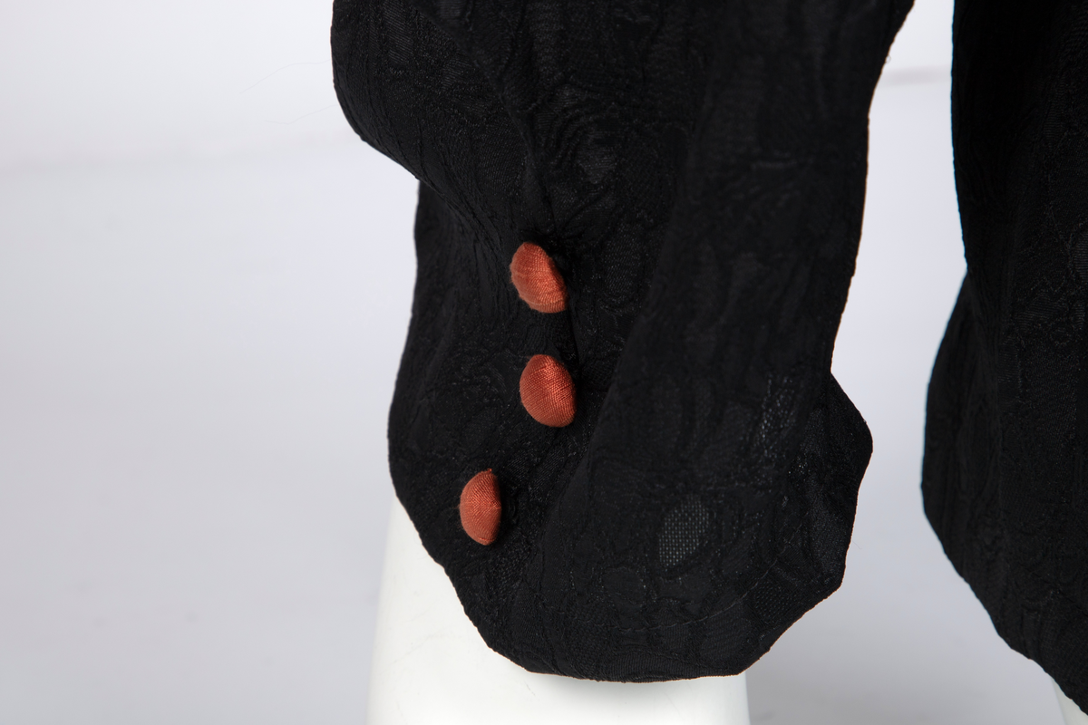 Ensemble bestående av kåpe, overdel, haremsbukse, hatt og mansjetter. Lang kåpe i rustrød råsilke med draperte ermer og detaljer i svart fløyel. Bladlignende dekor av blått og gullfarget stoff ved høyre skulder. Ermeløs overdel i svart råsilke og fløyel med svart syntetisk fôr. Overdelen går i en spiss opp ved bysten og i en spiss ned ved hoftene. Smale stropper over skuldrene. Dekorblomst i blått, gull og rustrødt i navlehøyde midt foran. Haremsbukse i svart syntetisk mønstervevd stoff med knapper kledd i rustrød råsilke nederst. Asymmetrisk hatt i svart ull- og silkeblanding, rustrød råsilke og polyesterbånd på innsiden. Mansjetter i råsilke med bomullsfôr.