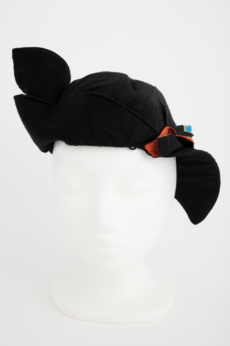 Ensemble bestående av kåpe, overdel, haremsbukse, hatt og mansjetter. Lang kåpe i rustrød råsilke med draperte ermer og detaljer i svart fløyel. Bladlignende dekor av blått og gullfarget stoff ved høyre skulder. Ermeløs overdel i svart råsilke og fløyel med svart syntetisk fôr. Overdelen går i en spiss opp ved bysten og i en spiss ned ved hoftene. Smale stropper over skuldrene. Dekorblomst i blått, gull og rustrødt i navlehøyde midt foran. Haremsbukse i svart syntetisk mønstervevd stoff med knapper kledd i rustrød råsilke nederst. Asymmetrisk hatt i svart ull- og silkeblanding, rustrød råsilke og polyesterbånd på innsiden. Mansjetter i råsilke med bomullsfôr.