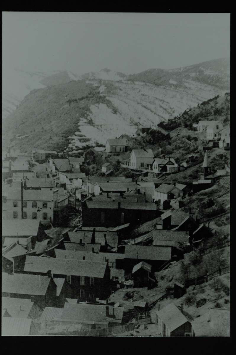 Et tettsted med flere bygninger i en dal. Åsside til høyre og fjell bak. Høyre halvpart av et større landskapsbilde.
