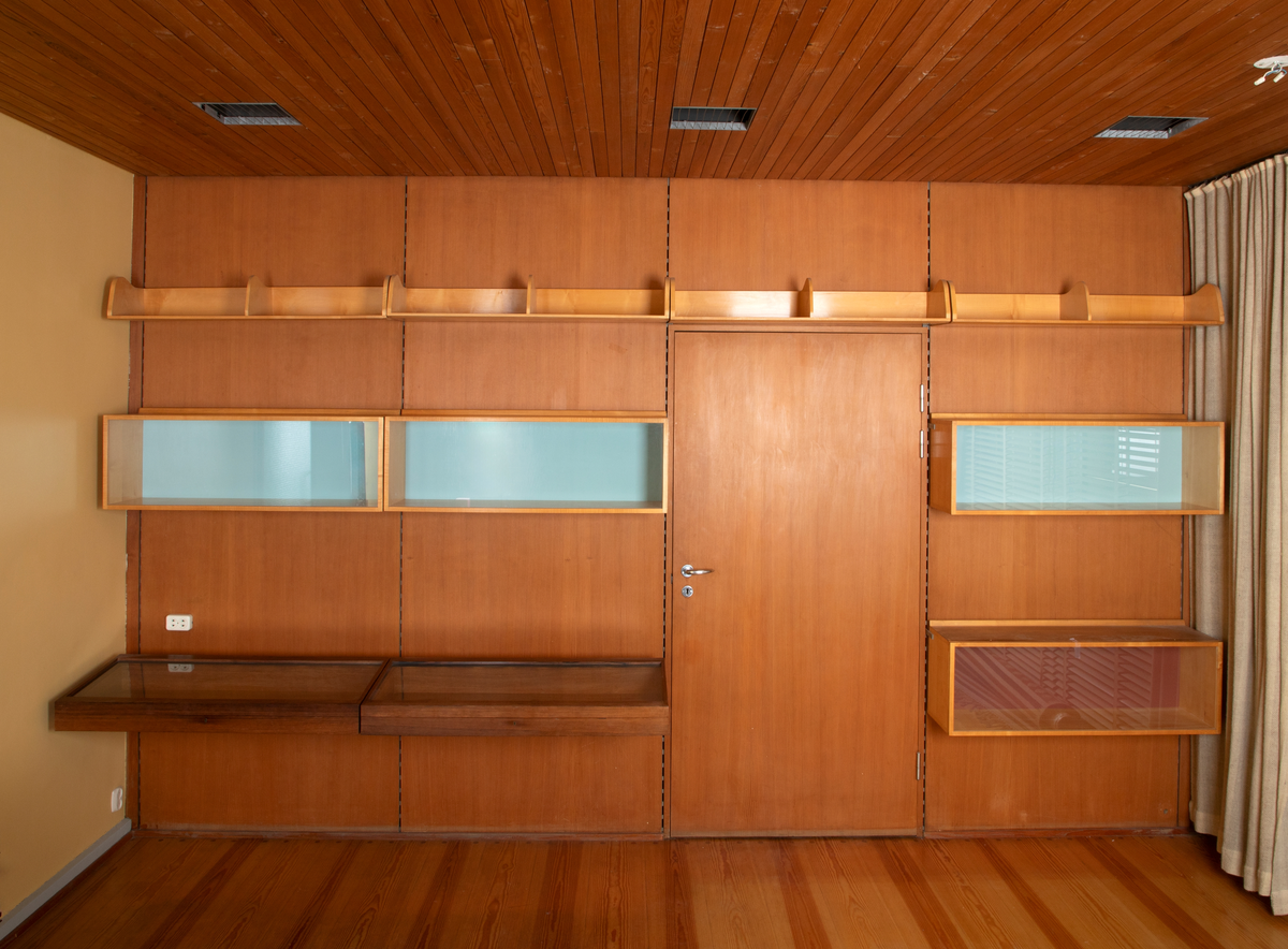 Panelvegg med flyttbare hylleseksjoner. Blant seksjonene finnes to diskmontre i mørkebrun rio-palisander med glass i lokket. Toppflatene skrån fremover, og lokkene er låsbare. Festet til bakvegg. Det er fire åpne reoler i dansk løn. Disse er to-det i bredden med lave skillevegger med buet profil. Det er fire lukkede veggmontre i dansk lønn. Glass på forsiden. Rektangulær kasseform. I tre av skapene er bakveggen malt lys blå, i den fjerde er bakveggen rødbrun. Bakveggen og dend rør er i douglas-gran