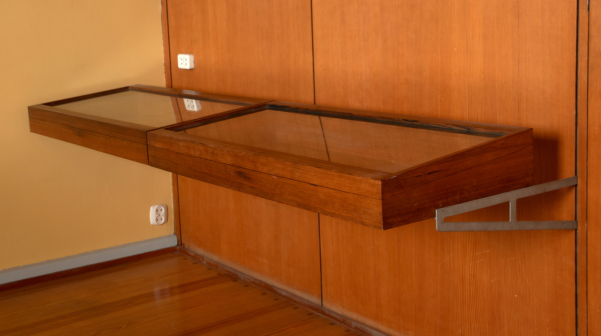 Panelvegg med flyttbare hylleseksjoner. Blant seksjonene finnes to diskmontre i mørkebrun rio-palisander med glass i lokket. Toppflatene skrån fremover, og lokkene er låsbare. Festet til bakvegg. Det er fire åpne reoler i dansk løn. Disse er to-det i bredden med lave skillevegger med buet profil. Det er fire lukkede veggmontre i dansk lønn. Glass på forsiden. Rektangulær kasseform. I tre av skapene er bakveggen malt lys blå, i den fjerde er bakveggen rødbrun. Bakveggen og dend rør er i douglas-gran