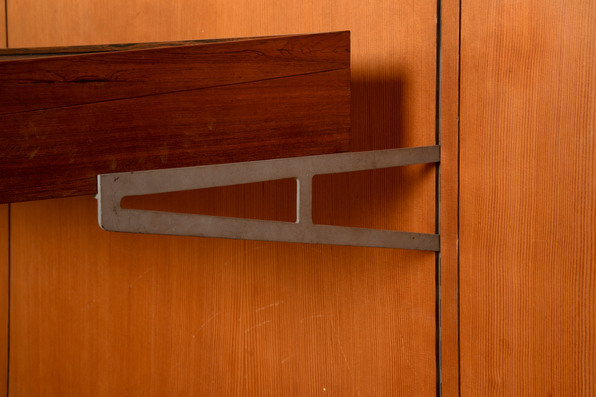 Panelvegg med flyttbare hylleseksjoner. Blant seksjonene finnes to diskmontre i mørkebrun rio-palisander med glass i lokket. Toppflatene skrån fremover, og lokkene er låsbare. Festet til bakvegg. Det er fire åpne reoler i dansk løn. Disse er to-det i bredden med lave skillevegger med buet profil. Det er fire lukkede veggmontre i dansk lønn. Glass på forsiden. Rektangulær kasseform. I tre av skapene er bakveggen malt lys blå, i den fjerde er bakveggen rødbrun. Bakveggen og dend rør er i douglas-gran