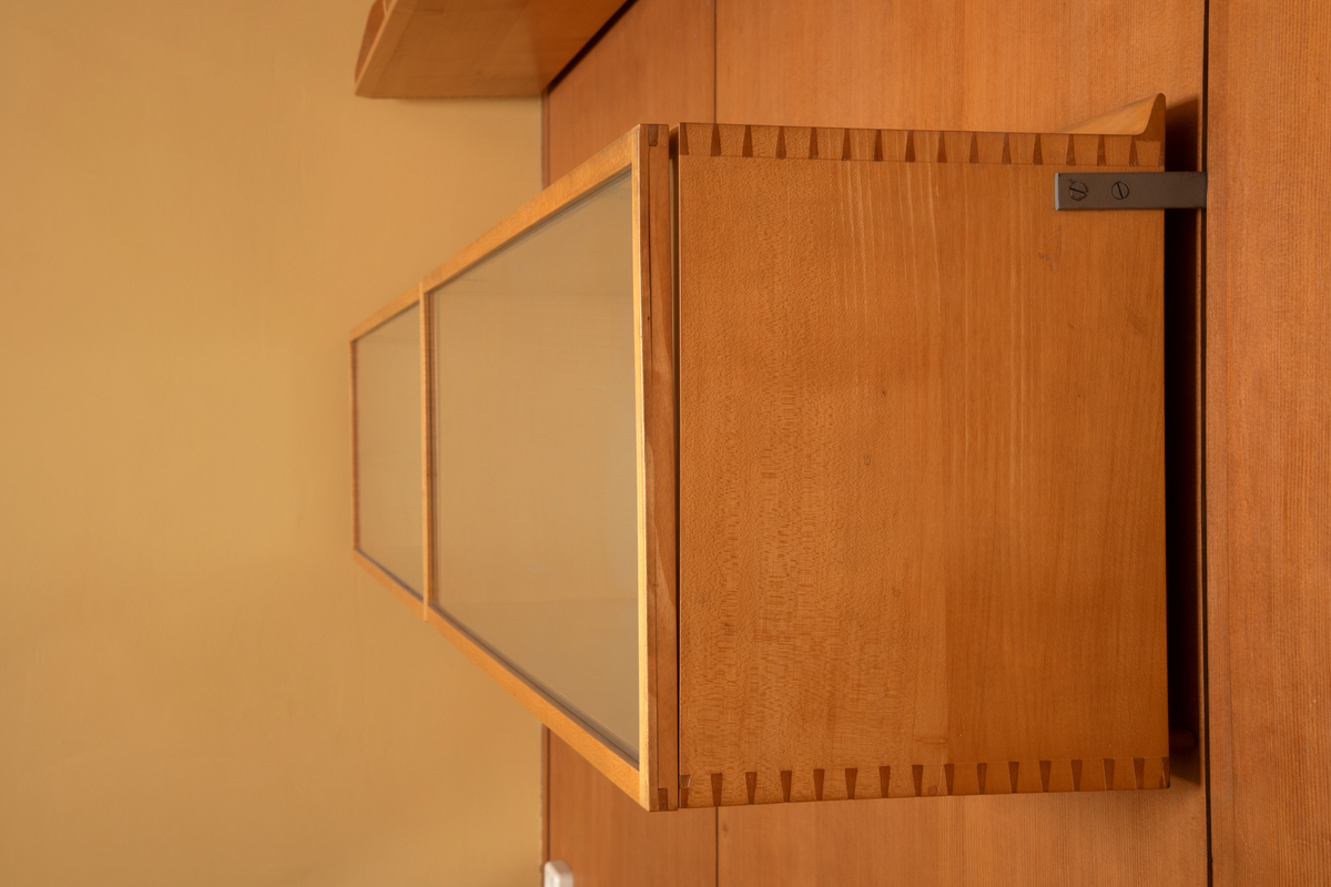 Panelvegg med flyttbare hylleseksjoner. Blant seksjonene finnes to diskmontre i mørkebrun rio-palisander med glass i lokket. Toppflatene skrån fremover, og lokkene er låsbare. Festet til bakvegg. Det er fire åpne reoler i dansk løn. Disse er to-det i bredden med lave skillevegger med buet profil. Det er fire lukkede veggmontre i dansk lønn. Glass på forsiden. Rektangulær kasseform. I tre av skapene er bakveggen malt lys blå, i den fjerde er bakveggen rødbrun. Bakveggen og dend rør er i douglas-gran