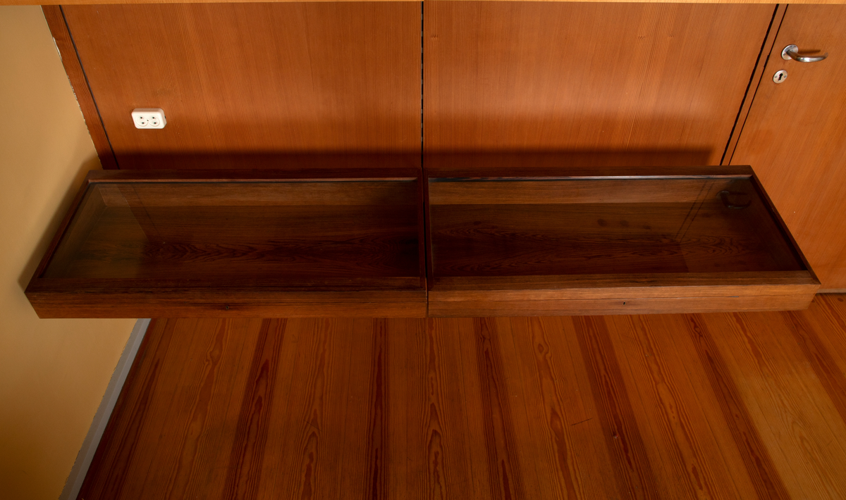 Panelvegg med flyttbare hylleseksjoner. Blant seksjonene finnes to diskmontre i mørkebrun rio-palisander med glass i lokket. Toppflatene skrån fremover, og lokkene er låsbare. Festet til bakvegg. Det er fire åpne reoler i dansk løn. Disse er to-det i bredden med lave skillevegger med buet profil. Det er fire lukkede veggmontre i dansk lønn. Glass på forsiden. Rektangulær kasseform. I tre av skapene er bakveggen malt lys blå, i den fjerde er bakveggen rødbrun. Bakveggen og dend rør er i douglas-gran