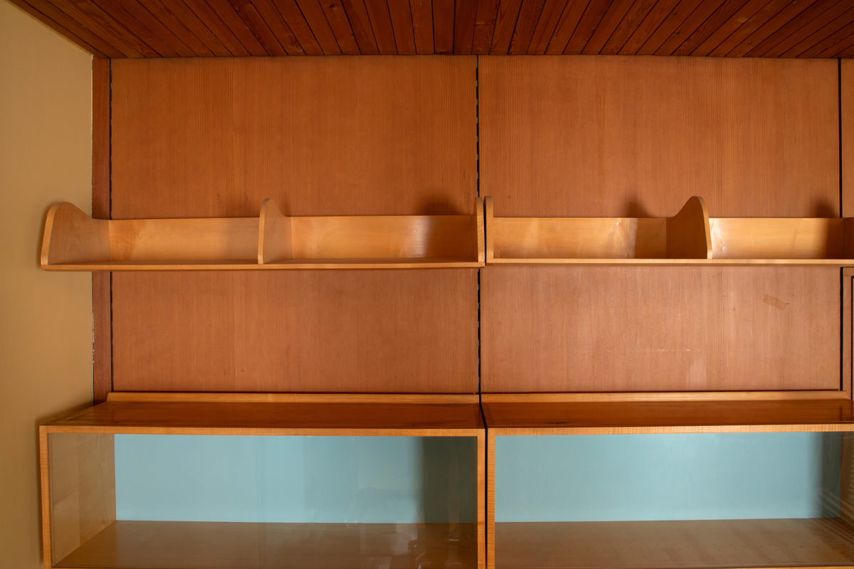 Panelvegg med flyttbare hylleseksjoner. Blant seksjonene finnes to diskmontre i mørkebrun rio-palisander med glass i lokket. Toppflatene skrån fremover, og lokkene er låsbare. Festet til bakvegg. Det er fire åpne reoler i dansk løn. Disse er to-det i bredden med lave skillevegger med buet profil. Det er fire lukkede veggmontre i dansk lønn. Glass på forsiden. Rektangulær kasseform. I tre av skapene er bakveggen malt lys blå, i den fjerde er bakveggen rødbrun. Bakveggen og dend rør er i douglas-gran