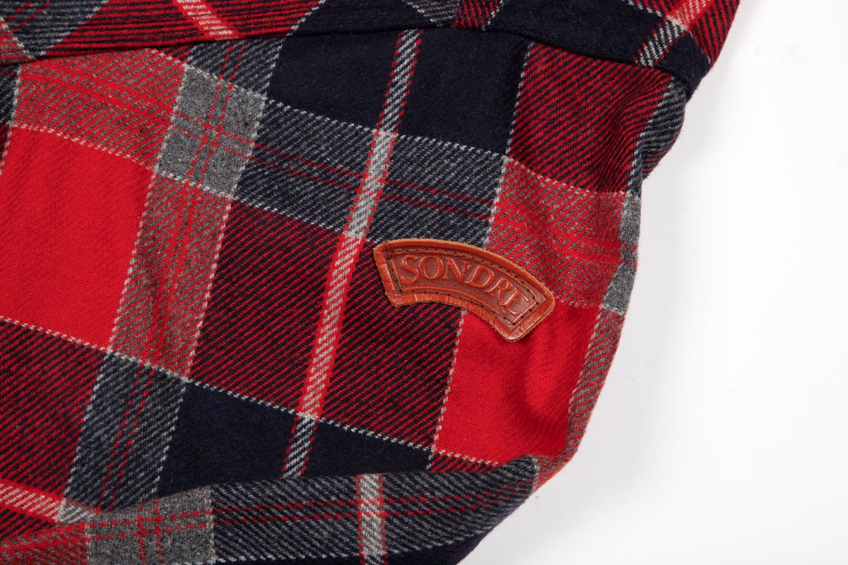 Rutete skjorte i ullblanding. Søm i hver side, litt oppsplittet nederst. Åpen foran med stolpe, knappet med seks knapphull og knapper. Dobbelt krage med små spisser og med brunt reinskinn på linningens utside. Bærestykke over skuldrene. En stor lomme på hvert forstykke. Et ekstra forstykke fra bærestykket foran danner klaffer med knapping for lommen. Skjorten er lett avrundet nedentil. Isydde ermer med mansjett og knapping. På albuene en skinnlapp og på venstre erme påsydd skinnlapp, hvorpå SONDRE.