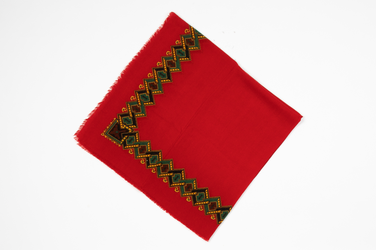 Sjal av rødt ullstoff, oppfrynset i kantene. Påtrykt mønsterbord i gult, grønt, brunt og mørkeblått. Mønsteret består av ruter tredd i hverandre med ornamenter i rutene.