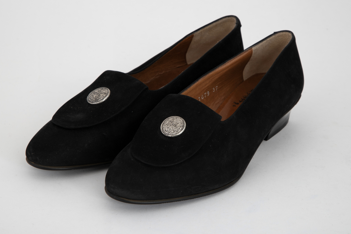 Sorte pumps i semsket skinn med lav hæl og rundet tå. Fremoverbøyet pløse med tinnknapp, knappen med gravert dekor. Fôret med brunt skinn. Sort, mønstret gummisåle.