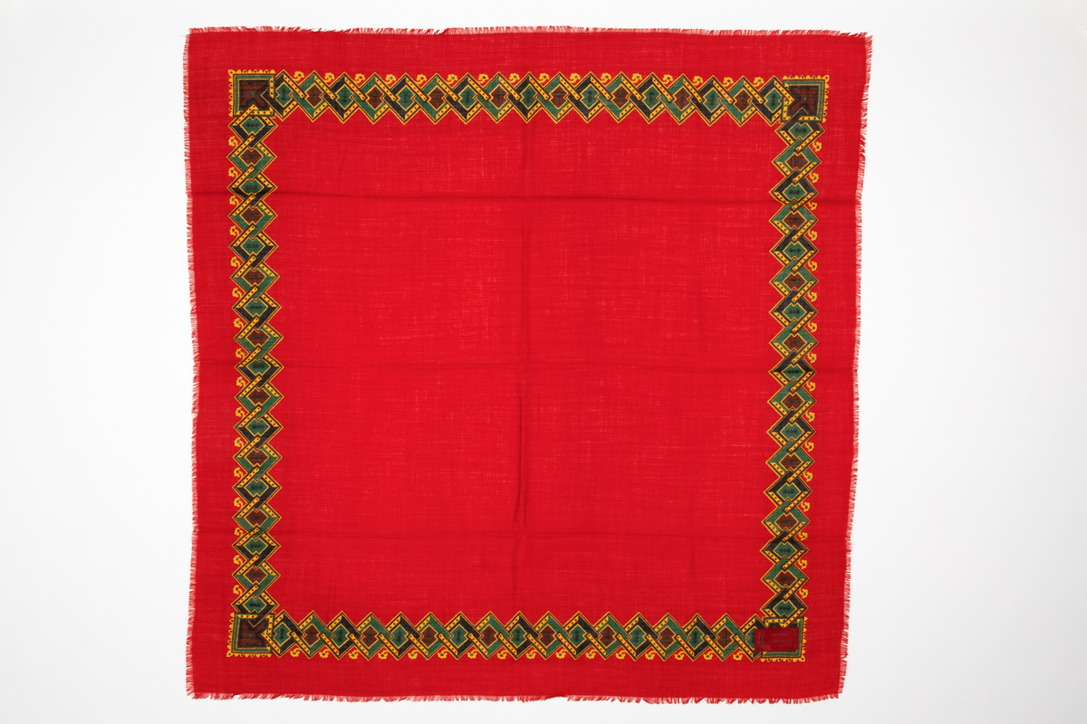 Sjal av rødt ullstoff, oppfrynset i kantene. Påtrykt mønsterbord i gult, grønt, brunt og mørkeblått. Mønsteret består av ruter tredd i hverandre med ornamenter i rutene.