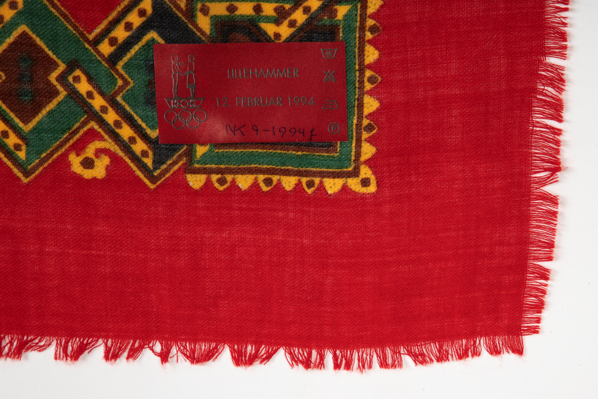 Sjal av rødt ullstoff, oppfrynset i kantene. Påtrykt mønsterbord i gult, grønt, brunt og mørkeblått. Mønsteret består av ruter tredd i hverandre med ornamenter i rutene.