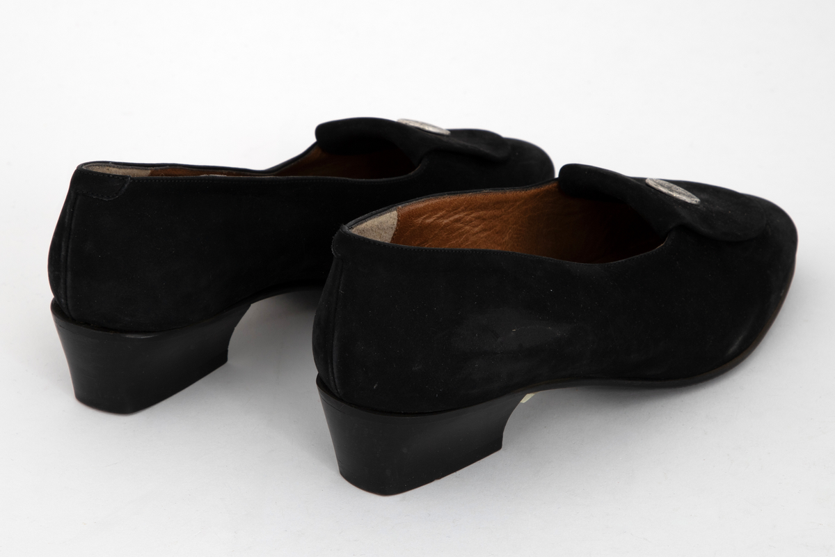 Sorte pumps i semsket skinn med lav hæl og rundet tå. Fremoverbøyet pløse med tinnknapp, knappen med gravert dekor. Fôret med brunt skinn. Sort, mønstret gummisåle.