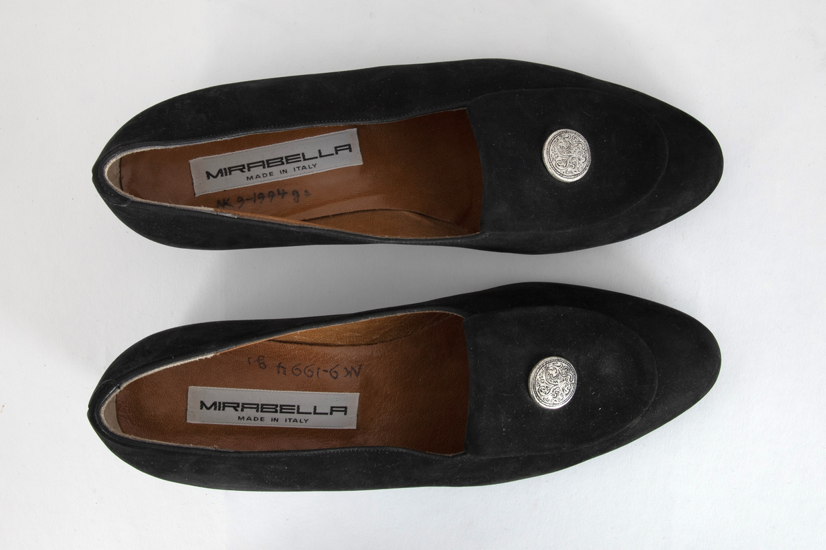 Sorte pumps i semsket skinn med lav hæl og rundet tå. Fremoverbøyet pløse med tinnknapp, knappen med gravert dekor. Fôret med brunt skinn. Sort, mønstret gummisåle.