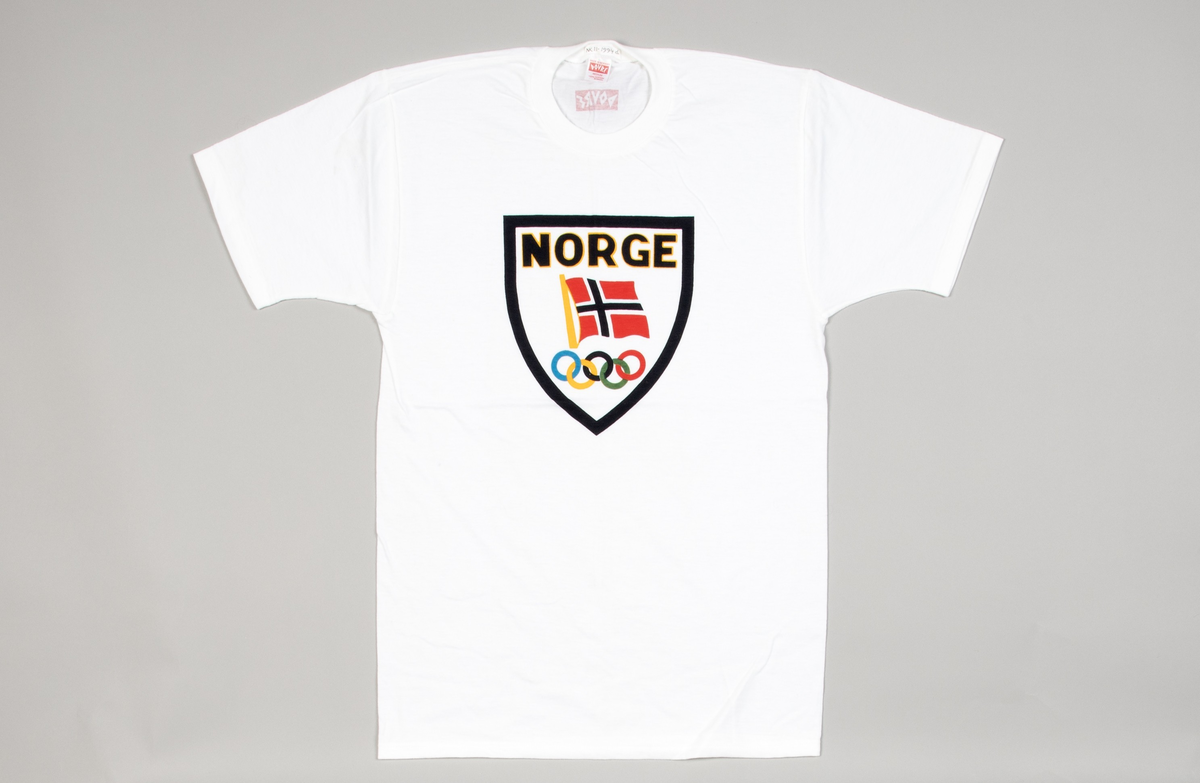 Kortermet t-skjorte i hvit bomullsjersey. Isydde ermer og rund vrangbordhals. På forsiden påtrykt innenfor sort skjoldformet ramme: NORGE i sort og gult, norsk flagg samt de olympiske ringer i sine forskjellige farger. Bak på nakken: DOVRE i hvitt på rød bunn.