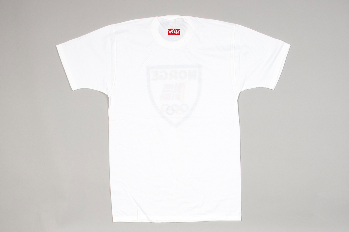 Kortermet t-skjorte i hvit bomullsjersey. Isydde ermer og rund vrangbordhals. På forsiden påtrykt innenfor sort skjoldformet ramme: NORGE i sort og gult, norsk flagg samt de olympiske ringer i sine forskjellige farger. Bak på nakken: DOVRE i hvitt på rød bunn.