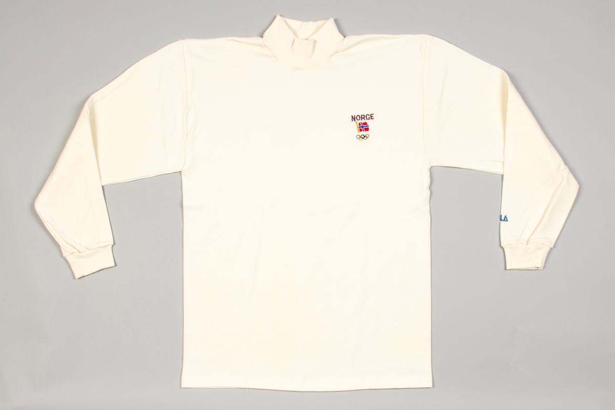 Langermet trøye av benhvit bomullsjersey med skuldersømmer. Rundstrikket bol og høyhalset, påsydd dobbelt hals. Isydde lange ermer med en søm og med påsydd dobbeltstrikket mansjett ved håndleddet. På bryst maskinbrodert: NORGE, norsk flagg og de olympiske ringer. På venstre erme i blått: AKLA.