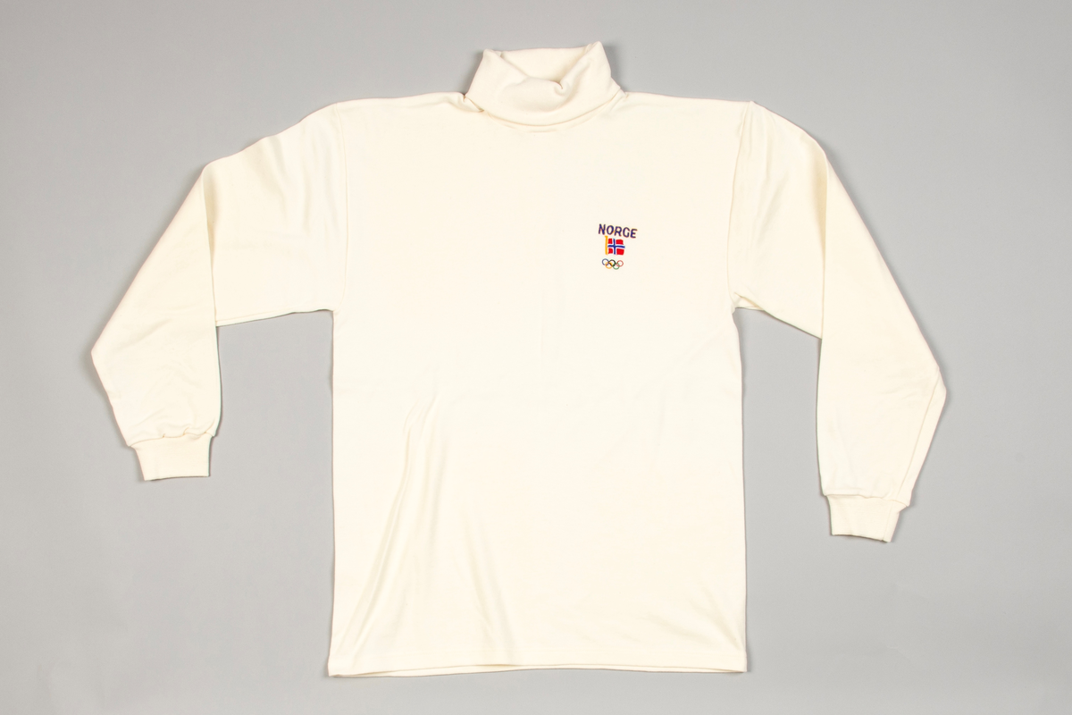 Langermet trøye av benhvit bomullsjersey med skuldersømmer. Rundstrikket bol og høyhalset, påsydd dobbelt hals. Isydde lange ermer med en søm og med påsydd dobbeltstrikket mansjett ved håndleddet. På bryst maskinbrodert: NORGE, norsk flagg og de olympiske ringer. På venstre erme i blått: AKLA.
