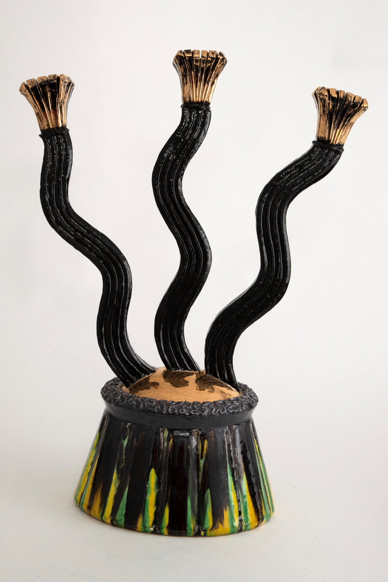 Trearmet kandelaber i keramikk. Fra en fot med rette, innskrånende vegger "vokser" tre lange, bølgende armer oppover. Armene ender i lyseholdere som øverst utvider seg og avsluttes med kvadratiske toppflater. Øverst på foten en kraftig rand som på toppen er teksturert. På fotens overside en buet flate som er glasert i gull. Fotens vegger, de tre armene og lyseholderne har en riflet overflate. Foten er glasert i sort med gule og grønne vertikale striper. Armene er sortglaserte, lysholderne er glasert i gull.