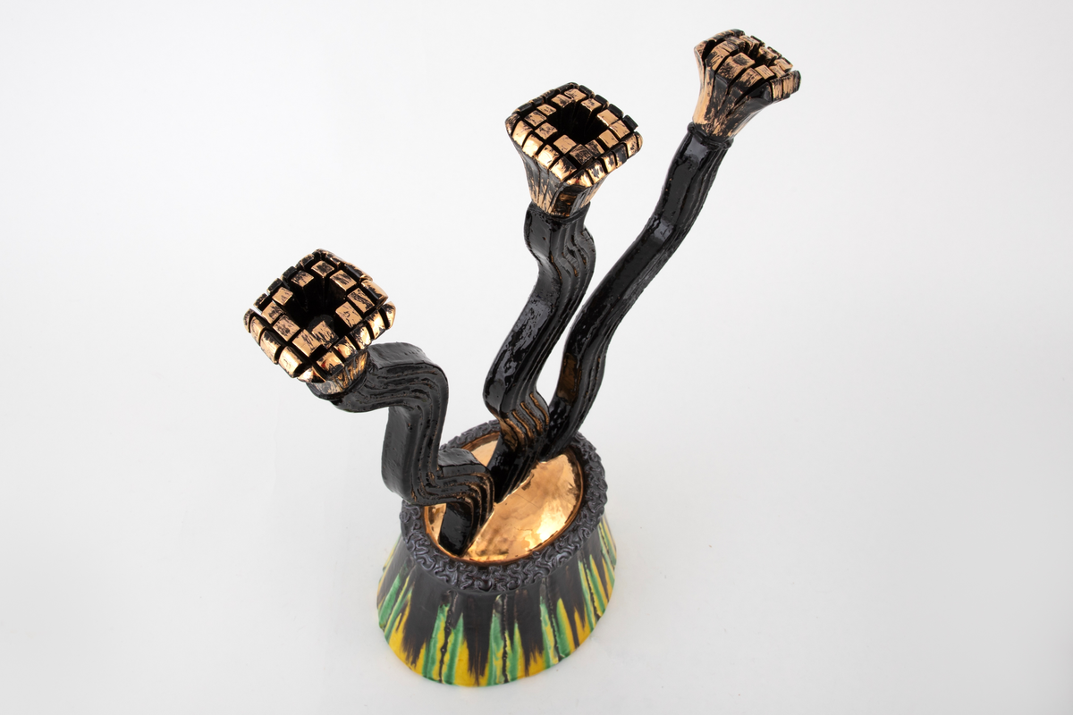 Trearmet kandelaber i keramikk. Fra en fot med rette, innskrånende vegger "vokser" tre lange, bølgende armer oppover. Armene ender i lyseholdere som øverst utvider seg og avsluttes med kvadratiske toppflater. Øverst på foten en kraftig rand som på toppen er teksturert. På fotens overside en buet flate som er glasert i gull. Fotens vegger, de tre armene og lyseholderne har en riflet overflate. Foten er glasert i sort med gule og grønne vertikale striper. Armene er sortglaserte, lysholderne er glasert i gull.