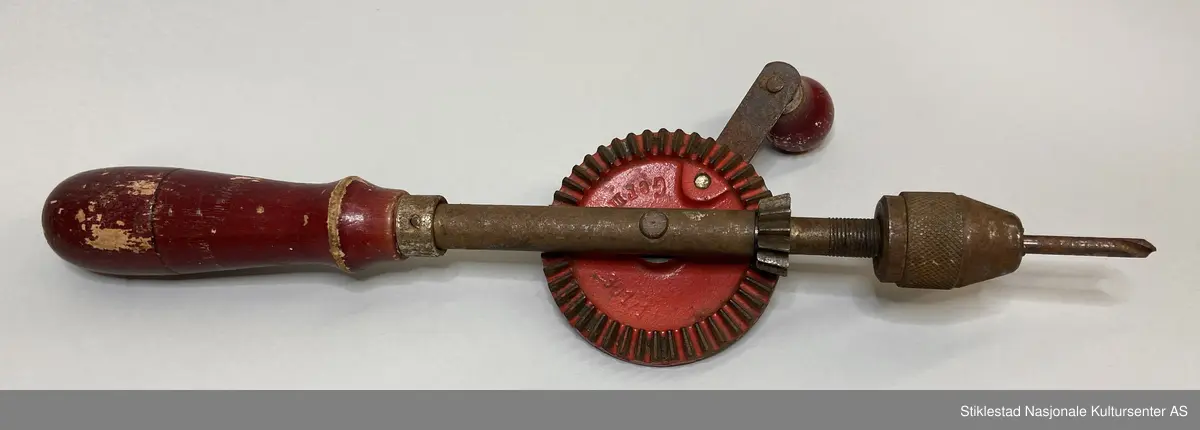 Fabrikkprodusert drill/hånddrill med rødfarget treskaft, sveiv og tannhjul. Ellers jern/metall. Det står en kort bor i chuck (kjoks). Merket 301/1 Germany på tannhjul/drivhjul. Andre siden et merke i gullfarge med rød skrift: Genko, Germany.