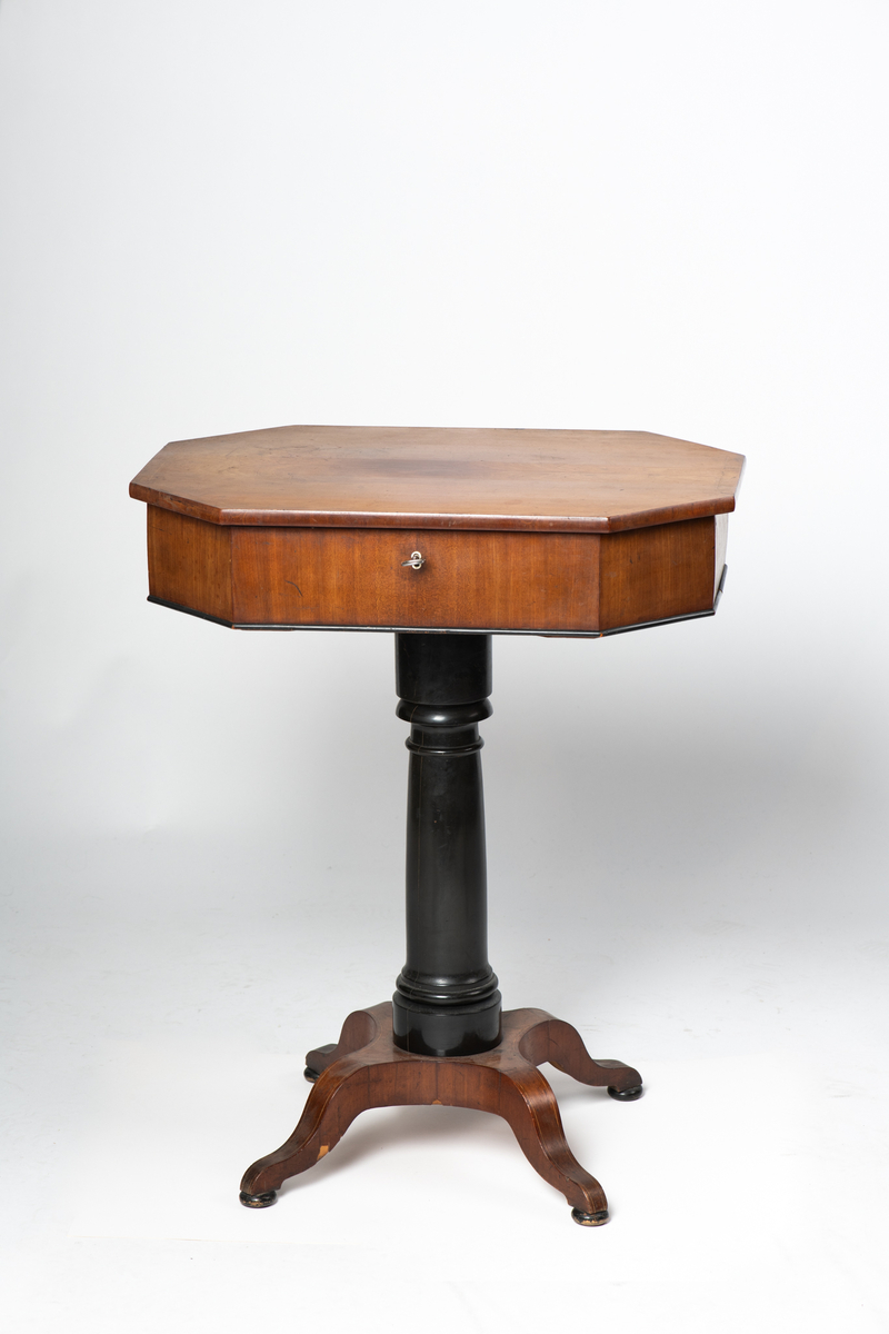 Søyle på firebenet fot. Åttekantet plate med en innlagt stripe med sitrontre langs kanten. Lite nøkkelskilt i sargen. Under platen inndelt i rom med løse lokk. Disse har  innlagte kantstriper og benknotter.
