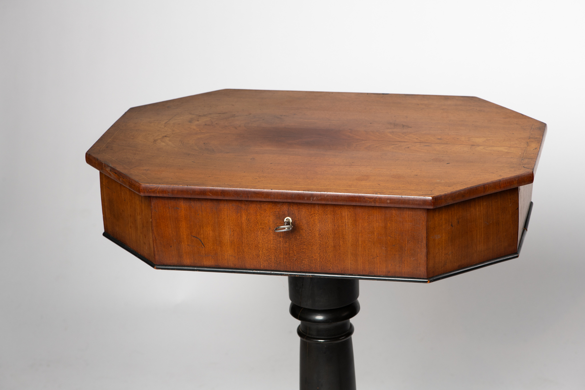 Søyle på firebenet fot. Åttekantet plate med en innlagt stripe med sitrontre langs kanten. Lite nøkkelskilt i sargen. Under platen inndelt i rom med løse lokk. Disse har  innlagte kantstriper og benknotter.
