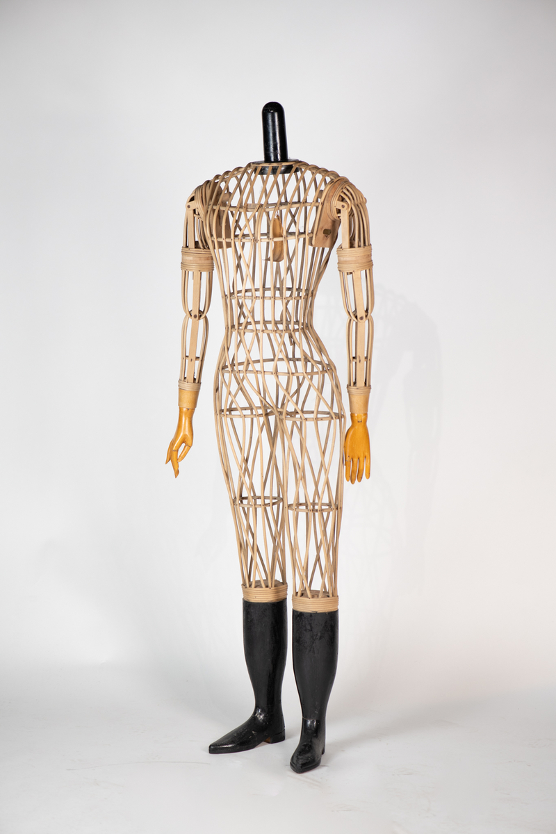 Skjelett  av trespiler, sortmalte  treføtter, lysbrune hender av tre. Kan sette på hode av voks. Skjelett med innsvunget talje, bevegelige armer, massive  føtter. Original etikett henger inni dukken.