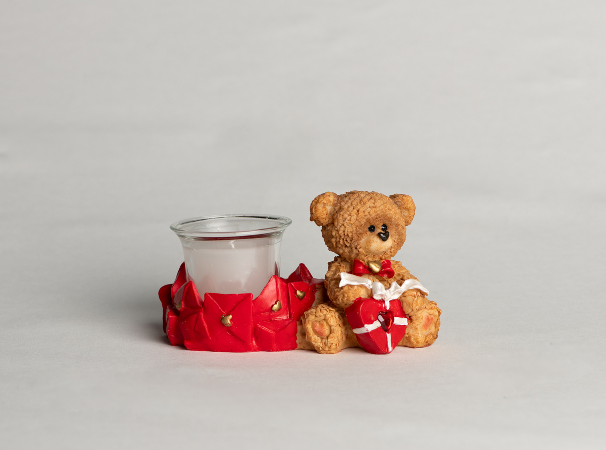 Bamse med sløyfe og hjerte rundt halsen, sittende, holder et hjerte med sløyfe i fanget. Ved siden av ligger mange uåpnede brev forseglet med hjerter, disse danner rundt fundament for det løse telysglasset.