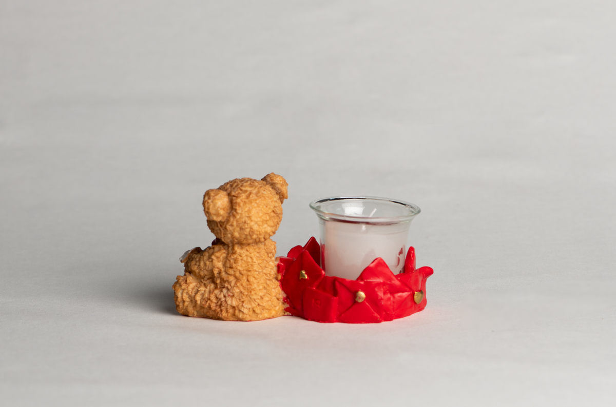 Bamse med sløyfe og hjerte rundt halsen, sittende, holder et hjerte med sløyfe i fanget. Ved siden av ligger mange uåpnede brev forseglet med hjerter, disse danner rundt fundament for det løse telysglasset.