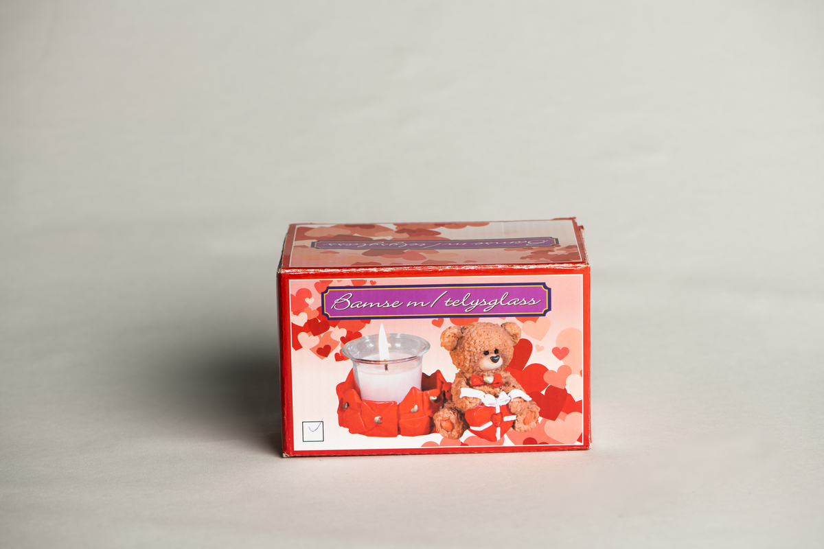 Bamse med sløyfe og hjerte rundt halsen, sittende, holder et hjerte med sløyfe i fanget. Ved siden av ligger mange uåpnede brev forseglet med hjerter, disse danner rundt fundament for det løse telysglasset.