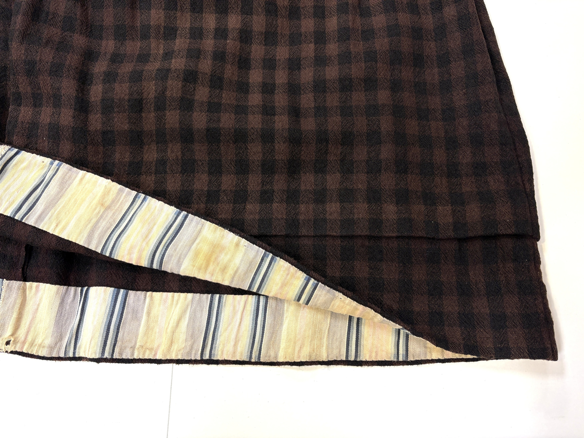 Stakk i samme stoff som liv SM.008023.01; svart og brunrutet ull i hjemmevevd toskaft. Stakken er falmet og slitt med en del hull spredt utover. Sydd sammen for hånd med attersting med svart bomullstråd av fem vevbredder som har jarekanter. Stakken har et 22 cm bredt glatt stykke midt på foran og foldelagt på begge sider. Åpningen er i venstre side og er 21 cm lang. Stakken har fire folder foldelagt mot høyre og seks folder foldelagt mot venstre, 3,5-4,5 cm brede. Midt på bak er det et 13 cm rynket felt med en halv fold over på begge sider. Foldene er tråklet med mørk blå ulltråd, sydd på med attersting med svart bomullstråd og så kastet med faldesting med svart bomullstråd. Linningen er i samme stoff som kjolen og sydd med faldesting med svart bomullstråd deretter med prikkesting med brun lintråd midt på linningen. Lukkes med to metallhekter. Stakken har en 4 cm bred hengefald 11,5 cm fra nedkanten, sydd med faldesting med svart bomullstråd, og en 7,5 cm bred skoning i stripet bomullsstoff sydd med faldesting med svart bomullstråd. Dette er kjøpetøy i fargene beige, rosa, lys blå, blå, mørk blå, hvit og grå. Stakken har en 21 cm lang og 19 cm bred lomme på høyre side i samme stoff som fôret på livet, sydd fast med attersting med ubleket bomullstråd. Stakken har en metallhempe midt på bak på linningen for å hekte på livet og skal brukes utenpå livet.