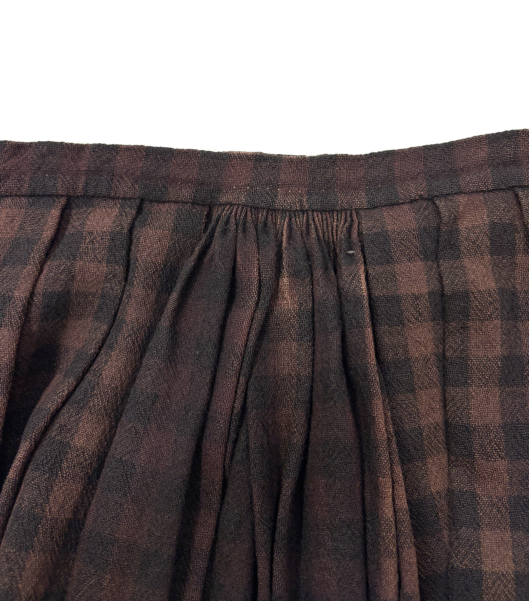 Stakk i samme stoff som liv SM.008023.01; svart og brunrutet ull i hjemmevevd toskaft. Stakken er falmet og slitt med en del hull spredt utover. Sydd sammen for hånd med attersting med svart bomullstråd av fem vevbredder som har jarekanter. Stakken har et 22 cm bredt glatt stykke midt på foran og foldelagt på begge sider. Åpningen er i venstre side og er 21 cm lang. Stakken har fire folder foldelagt mot høyre og seks folder foldelagt mot venstre, 3,5-4,5 cm brede. Midt på bak er det et 13 cm rynket felt med en halv fold over på begge sider. Foldene er tråklet med mørk blå ulltråd, sydd på med attersting med svart bomullstråd og så kastet med faldesting med svart bomullstråd. Linningen er i samme stoff som kjolen og sydd med faldesting med svart bomullstråd deretter med prikkesting med brun lintråd midt på linningen. Lukkes med to metallhekter. Stakken har en 4 cm bred hengefald 11,5 cm fra nedkanten, sydd med faldesting med svart bomullstråd, og en 7,5 cm bred skoning i stripet bomullsstoff sydd med faldesting med svart bomullstråd. Dette er kjøpetøy i fargene beige, rosa, lys blå, blå, mørk blå, hvit og grå. Stakken har en 21 cm lang og 19 cm bred lomme på høyre side i samme stoff som fôret på livet, sydd fast med attersting med ubleket bomullstråd. Stakken har en metallhempe midt på bak på linningen for å hekte på livet og skal brukes utenpå livet.